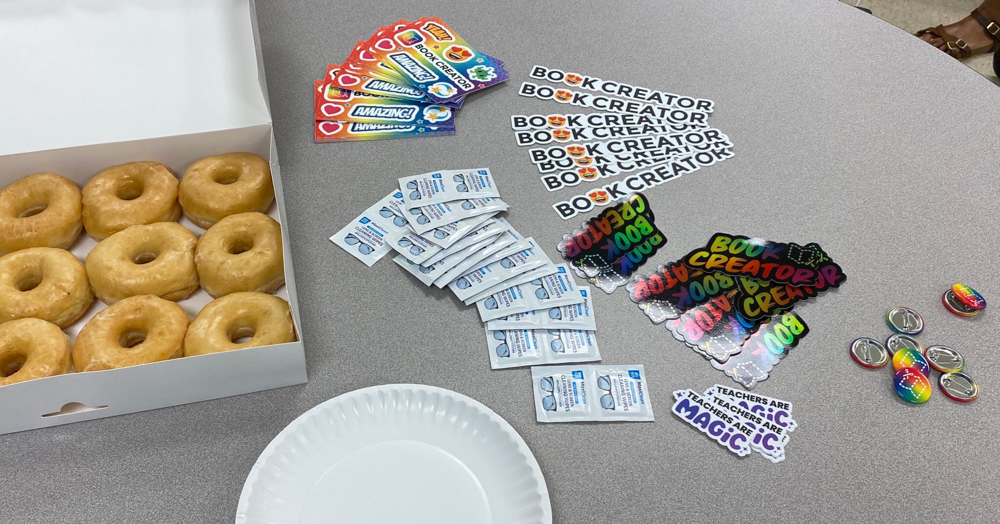 A table with a box of glazed donuts, packets of lens wipes, and colorful Book Creator-themed stickers and pins. The stickers include phrases like "Amazing" and "Book Creator," along with a few teacher-themed badges.