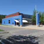 The image depicts Triton Regional High School, a modern building with a combination of brick and bright blue exterior walls. The school's name is displayed prominently on the blue section near the entrance, which features a white, curved awning. The area in front of the school is neatly landscaped with trees, and the surrounding driveway and parking area are clearly visible. The sky is bright blue, suggesting a clear, sunny day, contributing to the inviting and well-maintained atmosphere of the school grounds.