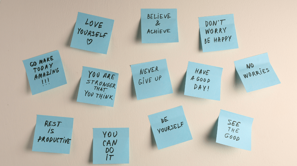 An arrangement of blue sticky notes on a beige wall, each featuring handwritten affirmations such as 'Love Yourself,' 'Believe & Achieve,' 'You Can Do It,' and 'Rest is Productive.' The notes radiate positivity and encouragement, promoting self-care and motivation.